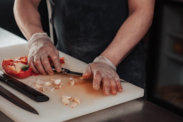 Hands crushing garlic and chopping red bell peppers on a white cutting board.